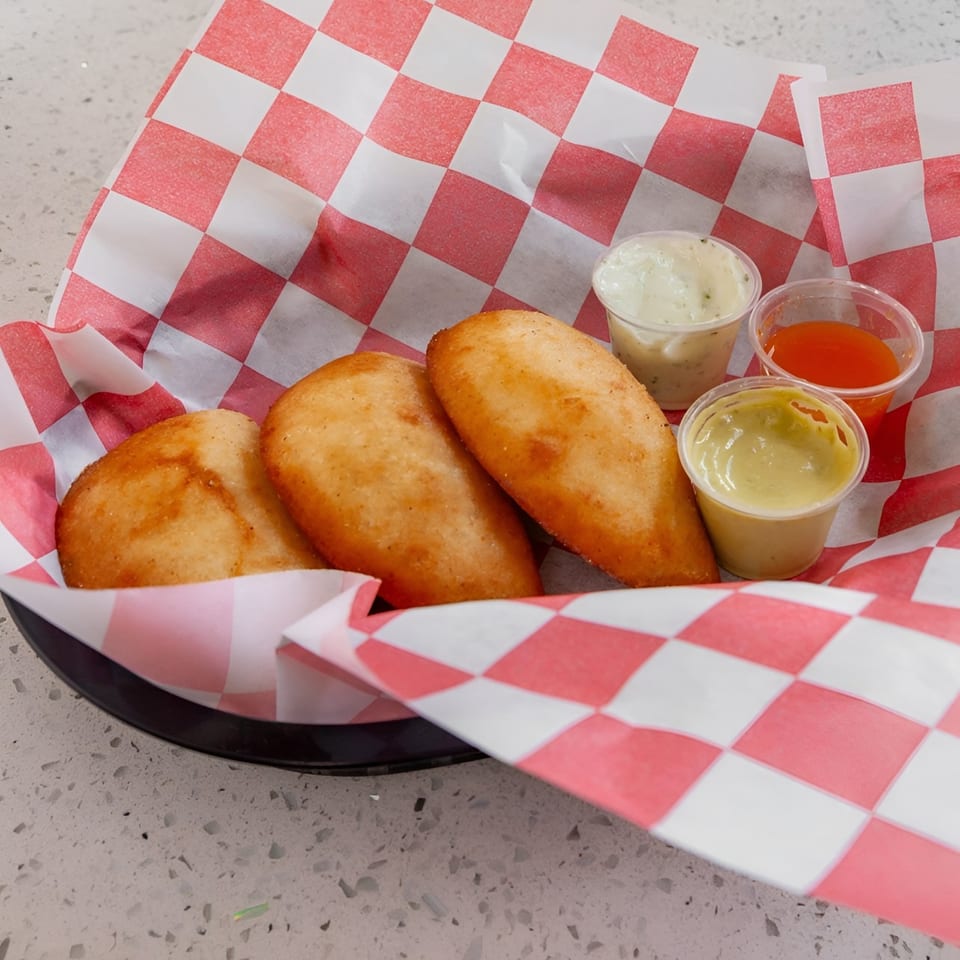 Three golden-brown empanadas in a basket lined with red and white checkered paper, with three dipping sauces.