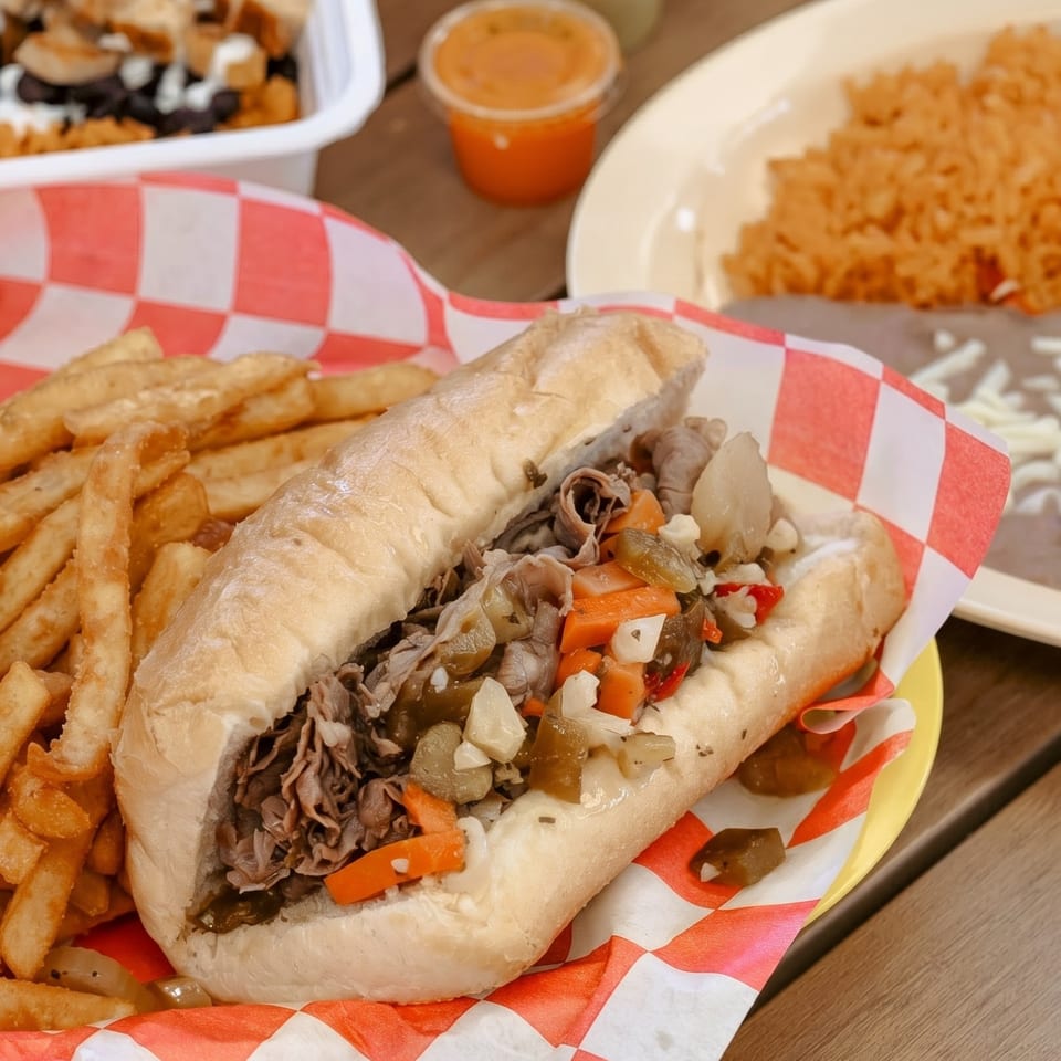 A Philly cheesesteak sandwich with fries in a red and white checkered basket, with rice and beans in the background.