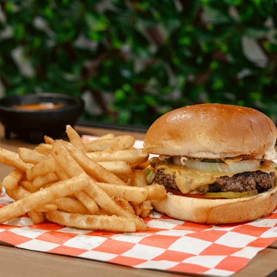 Cheeseburger with fries on red and white checkered paper, with a dark sauce bowl in the background.