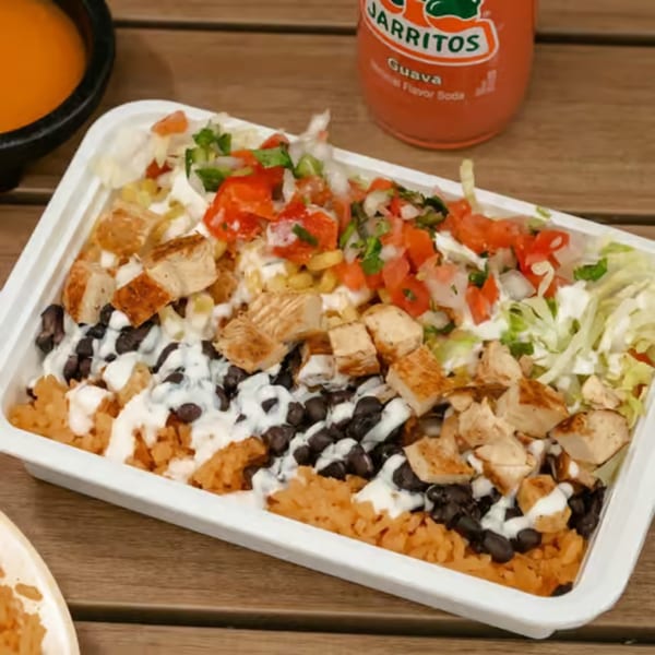 A white takeout container filled with chicken, rice, black beans, salsa, and lettuce, with a Jarritos soda.