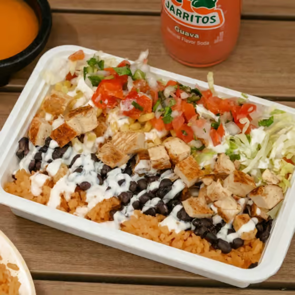 A white takeout container filled with chicken, rice, black beans, salsa, and lettuce, with a Jarritos soda.